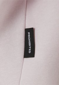 Tessuto rosa chiaro con un'etichetta nera che mostra la parola "PROIBITO" in testo bianco, cucita nel materiale di lato.
