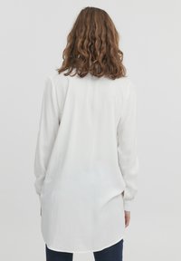 Langärmlige, weiße Bluse aus leichtem Stoff, mit abgerundetem Saum und einem subtilen Glanz. Rückansicht mit lockerer Passform.