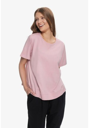 Młoda kobieta uśmiechająca się, ubrana w luźną różową koszulę z krótkim rękawem i czarne spodnie, z rękami w kieszeniach na białym tle.