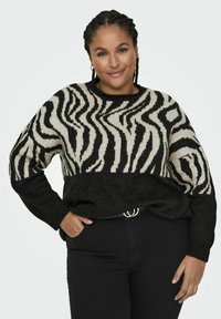 Črno in kremno zebraste pletene pulover s črnim spodnjim delom in rebrastimi manšeti, v kombinaciji s črnimi hlačami in srebrnim pasom.