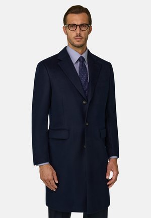 Mann mit schwarz gerahmter Brille, dunkelblauem Mantel, blau gemustertem Krawatte und gestreiftem Hemd vor einfarbigem Hintergrund.