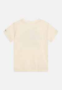 T-shirt beige clair à manches courtes en coton, avec un design simple, un col rond et un petit logo sur la manche gauche.