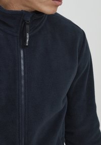 Chaqueta de forro polar azul marino con cremallera, con un cuello alto, que presenta una cremallera de contraste y una etiqueta que dice "11 PROJECT" en el tirador de la cremallera.