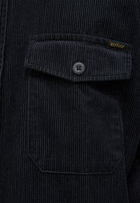 Marineblaue Cordjacke mit einer Brusttasche mit Knopf und einem kleinen gelben Markenschild. Texturiertes, geripptes Muster auf dem Stoff.