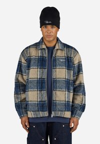 Fleece zip jacket featuring a blue and beige plaid pattern, featuring a collar and ribbed cuffs. Worn over a navy shirt and paired with jeans.