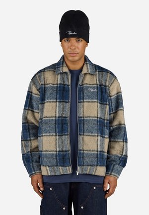 Fleece zip jacket featuring a blue and beige plaid pattern, featuring a collar and ribbed cuffs. Worn over a navy shirt and paired with jeans.