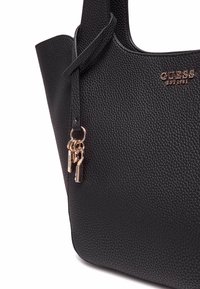 Bolso tote de cuero negro texturizado con detalles lisos y un logo de marca en tono dorado. Presenta una correa desmontable con hardware de llavero decorativo.