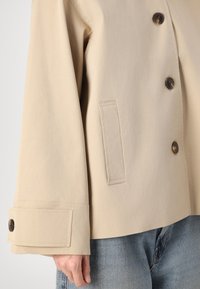 Beige oversize jakke med knaplukning, der har store knapper, en brystlomme og brede manchetter. Fremstillet af et letvægtsstof.