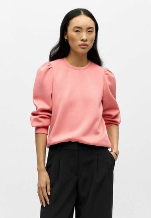 Sweatshirt rose à manches ballons, col rond et texture douce, associé à un pantalon noir ajusté. Design simple sans motifs visibles.