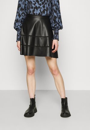 Femme portant une jupe en cuir noire, des bottines noires et un chemisier bleu à manches longues à motifs, debout devant un fond clair uni.
