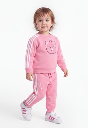 Peuter die een roze trainingspak draagt met een berengezicht op de trui en witte sneakers met roze strepen, staat en lacht.