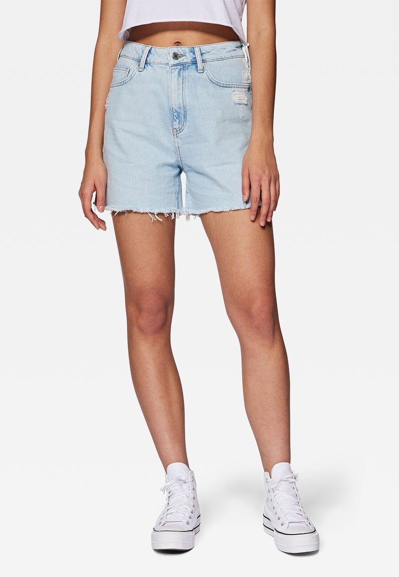Femme portant un short en denim bleu clair déchiré avec un ourlet brut, des baskets blanches montantes et un haut court blanc, debout devant un fond uni.