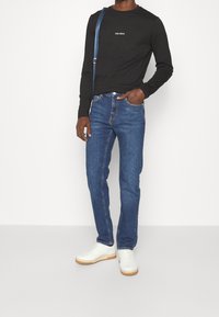 Schwarzes Langarmshirt mit "LES DEUX" Schriftzug, blaue Denim-Jeans, weiße Sneakers und ein blauer Umhängegurt quer über den Körper.