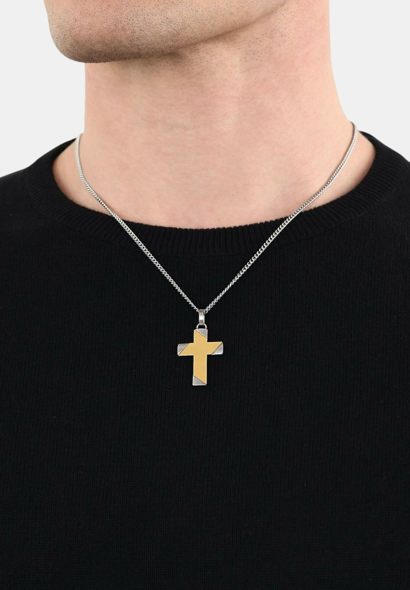 Zilveren ketting met een vergulde en zilveren kruispendant, met een slank ontwerp en een gladde textuur, gedragen over een zwarte trui.
