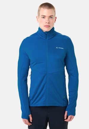 Jonge man draagt een blauw jack met ritssluiting en duimgaten, staat voor een effen lichte achtergrond en kijkt recht vooruit.