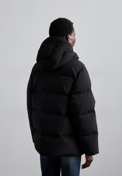 Black puffer jacket with a hood, featuring large quilted sections and a relaxed fit. Soft texture, suitable for cold weather. Jeans visible below.