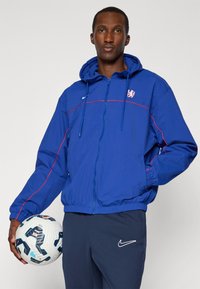 Blue jacket with a hood and red stripes, featuring a logo on the chest, paired with dark pants and a soccer ball. Smooth fabric texture.