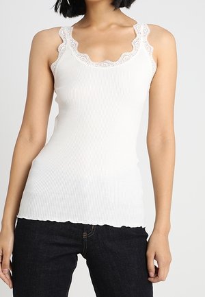 Mujer que lleva una camiseta de tirantes blanca acanalada con ribete de encaje y vaqueros negros, de pie frente a un fondo liso y claro.