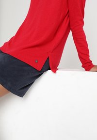Top rosso a maniche lunghe in maglia con texture a coste, dotato di uno spacco laterale e un accento con logo in metallo. Abbinato a pantaloni corti blu navy.
