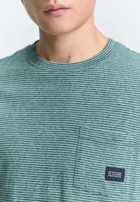 Groene gestreepte t-shirt met een ronde hals, heeft een borstzakje en een klein merketiket. Materiaal lijkt zacht met een gestructureerde afwerking.