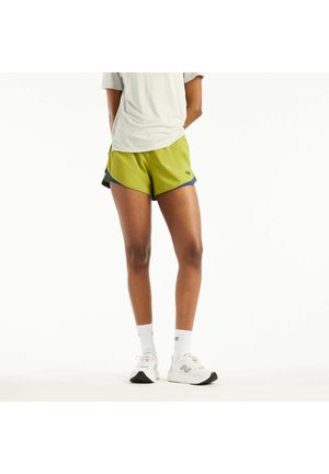 Person iført lysegrønne atletikshorts, hvide sokker og hvide New Balance-sneakers, der står med hænderne bag ryggen.
