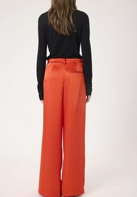 Pantalons larges en satin orange avec une texture lisse, dotés d'une ceinture et d'un détail de poche arrière. Vue de dos. Haut noir à manches longues.