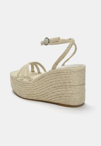 Sandale compensée en toile beige avec texture en corde tressée, bout ouvert et sangle de cheville réglable avec boucle métallique.