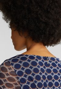 Mörkblå klänning med cirkulärt mönster i vitt och orange. Har en transparent meshsärmling och en detalj med bakdragkedja.