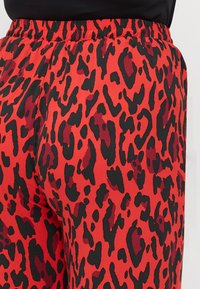 Kalhoty s červeným leopardím vzorem a černým elastickým pasem, které mají volný střih, zdůrazňující strukturovanou látku a odvážný kontrast vzorů.