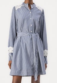 Randig blå och vit skjortklänning med långa ärmar, spetsdetaljer på axlarna, knäppning fram och knytbar midjebälte.