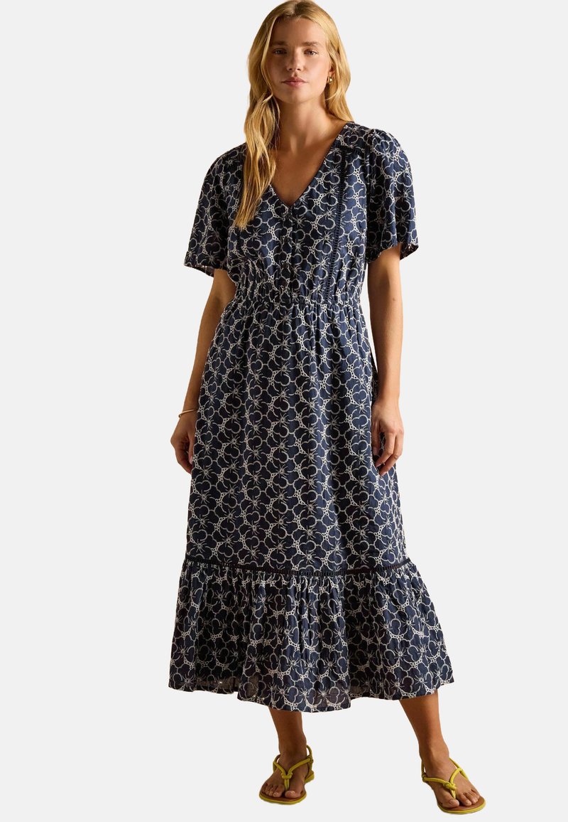 Marineblaue Maxikleid mit floralem Muster, kurzenÄrmeln, gerafftem Taillenbereich und rüschendem Saum. Getragen mit gelben Flip-Flops.