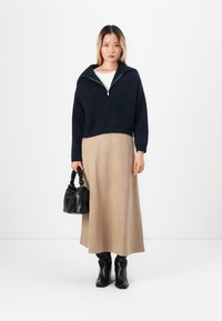 Kvinne iført en marineblå zip-up jakke, beige midiskjørt, svarte hælede støvler, holder en svart handbag mot en helt hvit bakgrunn.