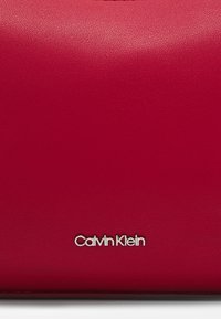 Borsa a tracolla in pelle rossa con una texture liscia e un logo Calvin Klein argentato. Presenta una forma rettangolare e un design minimalista.