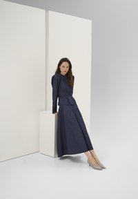 Vestito di denim blu navy con corpetto aderente, maniche a sbuffo e gonna lunga. Abbinato a tacchi a punta di colore grigio chiaro. Sfondo minimalista.