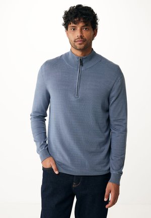 Pullover azzurro chiaro a maniche lunghe con colletto alto e chiusura con zip. Presenta una texture liscia e polsini a costine. Indossato con jeans scuri.