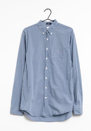 Chemise à manches longues boutonnée à petits carreaux bleus et blancs, avec poche avant, suspendue à un cintre noir sur fond blanc.