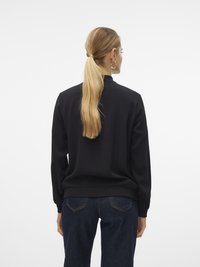 Ženska z dolgim svetlo blond čopom, oblečena v črn sweatshirt in temno modre kavbojke, prikazana zadaj na enobarvni beli podlagi.