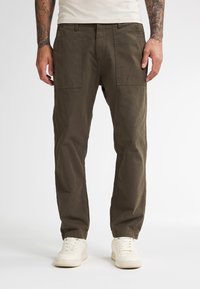 Uomo in pantaloni dritti marroni con ampie tasche anteriori e sneakers bianche, in piedi contro uno sfondo bianco uniforme.