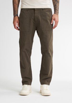 Homme portant un pantalon droit marron avec de grandes poches avant et des baskets blanches, se tenant devant un fond blanc uni.