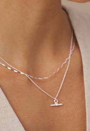 Deux délicats colliers en argent autour du cou d'une personne, l'un avec un petit pendentif barre horizontale et l'autre avec une fine chaîne en zigzag.