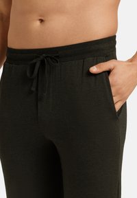 Tmavě zelené jogger kalhoty vyrobené z měkké tkaniny, s elastickým pasem se šňůrkou a bočními kapsami. Hladká textura.