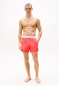 Pantaloni scurți de baie pentru bărbați, de culoare coral strălucitor, cu un tiv alb și un mic patch de marcă pe partea stângă. Material neted și ușor.