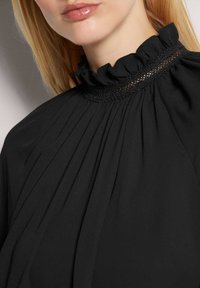 Schwarze Bluse mit hohem Kragen, verziert mit einer Rüsche, plissierter Stofftextur und feinen Spitzenverzierungen am Halsausschnitt.