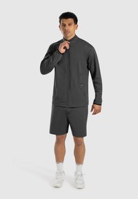 Dunkelgraue Zip-Jacke mit hohem Kragen, kombiniert mit passenden Shorts. Getragen mit weißen Sportschuhen und Socken. Glatte Textur, minimalistisches Design.