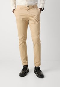 Beige katoenen chino's met een slim fit, voorzien van voorzakken en een enkele knoopsluiting, gecombineerd met zwarte leren veterschoenen.