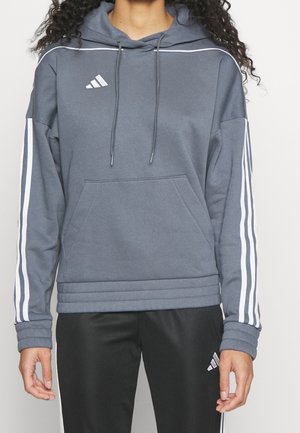 Persoon draagt een grijze Adidas-hoodie met witte strepen op de mouwen en zwarte Adidas-broek met witte strepen langs de zijkanten.