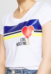 Bílé tričko s krátkým rukávem se modrými, žlutými a černými horizontálními pruhy. Obsahuje grafiku srdíčkového zmrzliny a nápis "LOVE MOSCHINO".