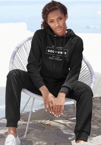 Schwarzes Fleece-Hoodie und Jogginganzug-Set mit dem Aufdruck "Good Vibes" in Weiß, mit gerippten Bündchen und einer verstellbaren Kapuze, vor einem hellen Hintergrund.