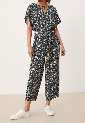 Vrouw draagt een zwart-witte jumpsuit met bloemenprint, een ceintuur in de taille en witte sneakers, staand tegen een effen achtergrond.