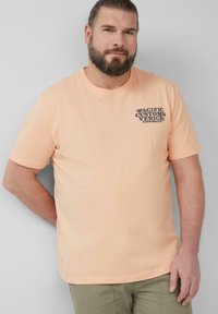 Ein bärtiger Mann trägt ein pfirsichfarbenes T-Shirt mit dem Aufdruck "Pacific Customs Venice" auf der Brust und steht vor einem hellgrauen Hintergrund.